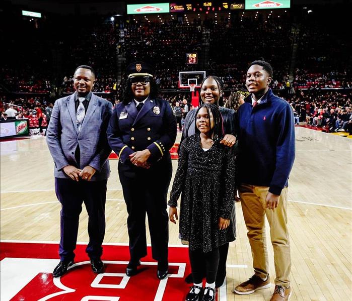 A group of five people, including a uniformed officer, stand together on a basketball court with a crowd in the background. 